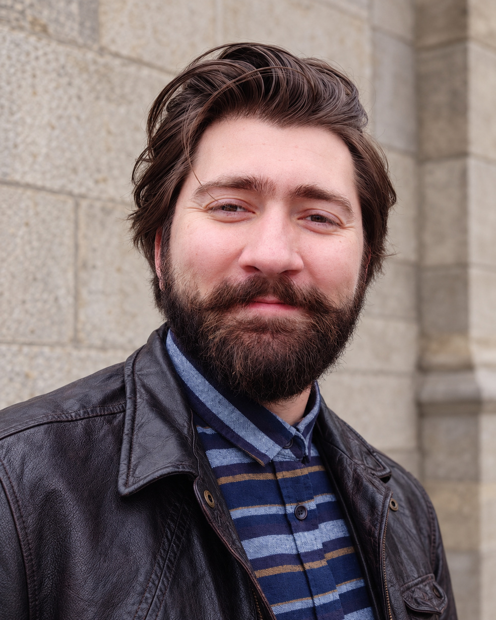 Rron Imami wearing a leather jacket and checked shirt against a stone background.